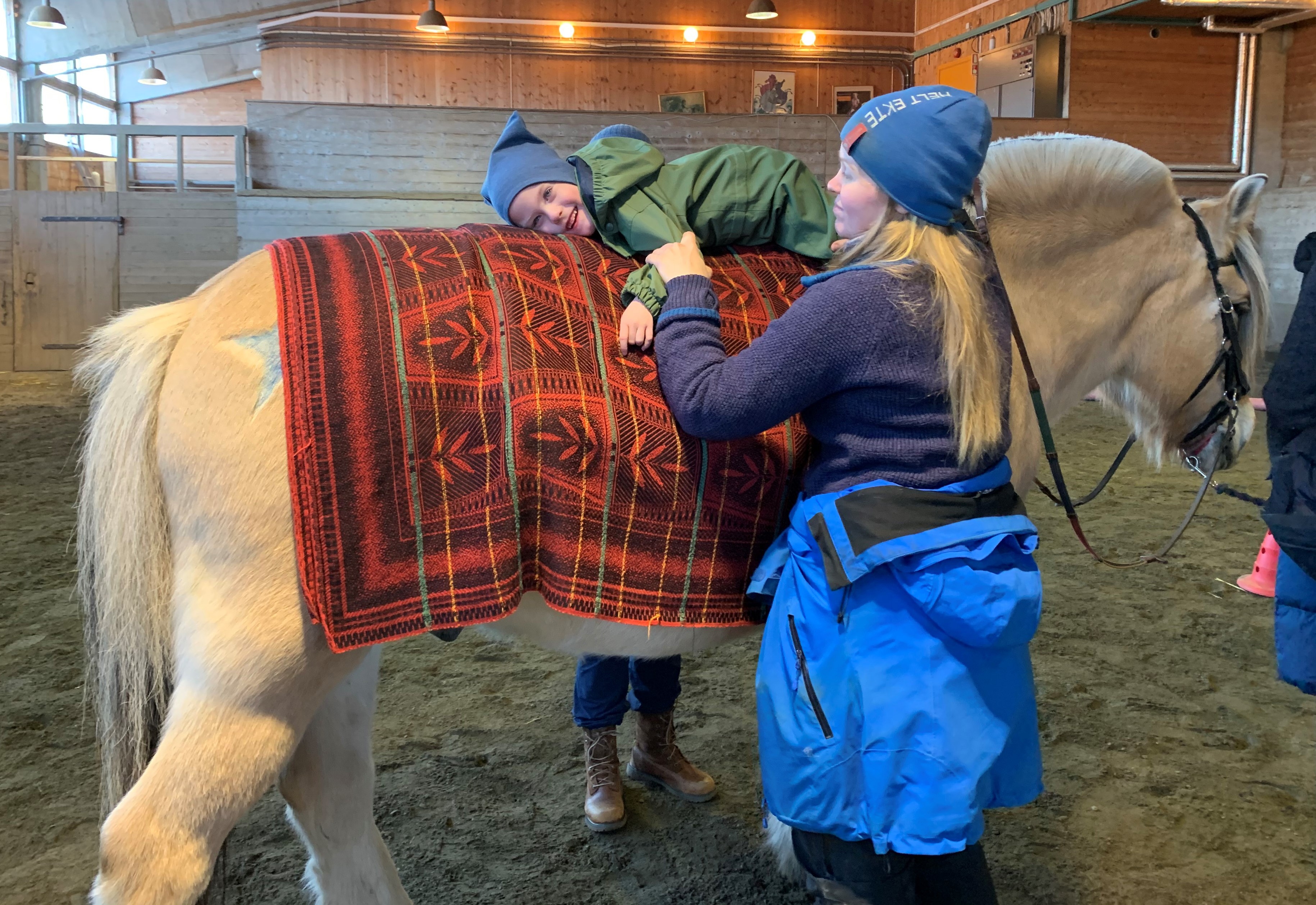 En smilende gutt ligger på magen på hesteryggen, mens rideinstruktøren holder han fast